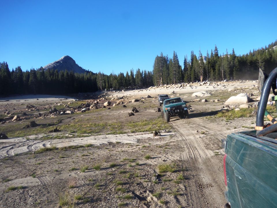 Green off-road vehicles on a dirt track with trees and a mountain in the background
