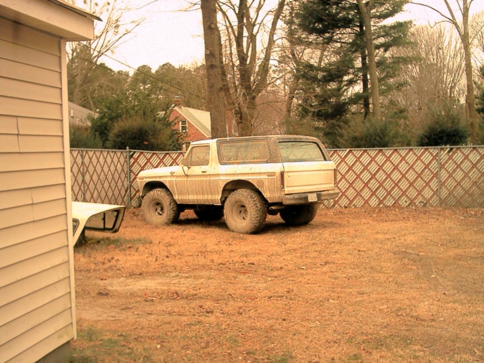 Muddy white Ford Bronco with large off-road tires parked in a backyard by a lattice fence, rear three-quarter view
