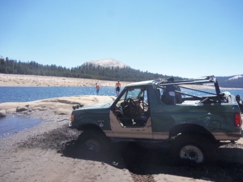 1968 Ford Bronco - An off-road SUV with a roll cage sits on the sandy edge of a lake under a clear blue sky.