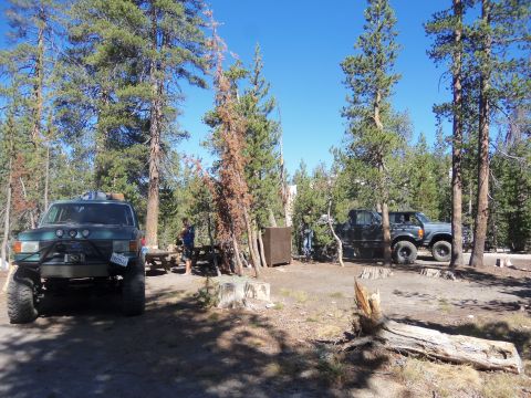 1968 Ford Bronco - Two off-road trucks are parked in a wooded campsite under a clear blue sky.