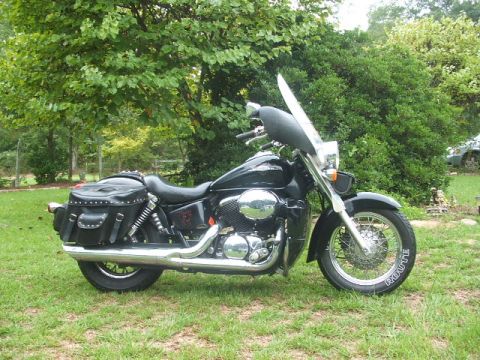 1998 Honda Shadow VT750C - A black cruiser motorcycle with saddlebags and a windshield is parked on a grassy lawn with trees in the background.