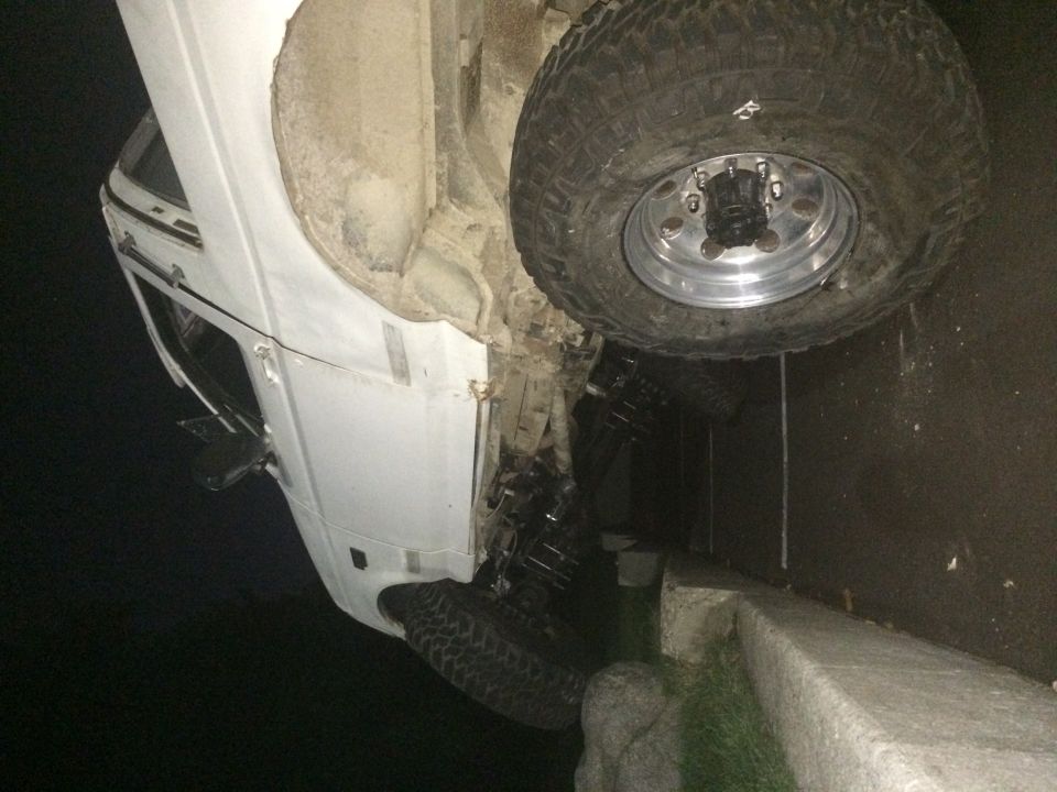 White pickup truck, possibly a Ford Bronco, tilted on its side. Large off-road tires are visible