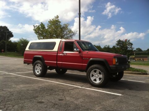 1991 Jeep Comanche - A red Jeep Comanche pickup truck with a white camper shell is parked in a lot under a cloudy sky.