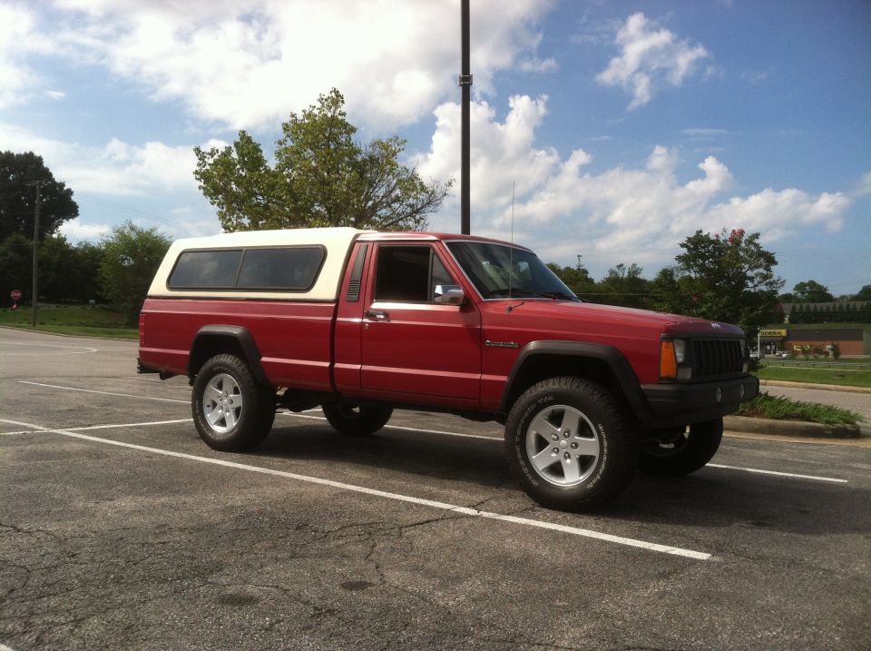 Red Jeep Comanche pickup with a white camper shell parked in a lot