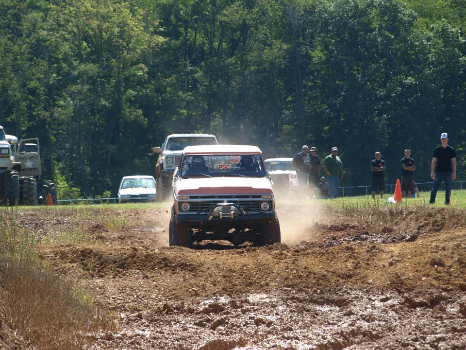 Off-road trucks driving through mud. One truck in foreground is orange and white
