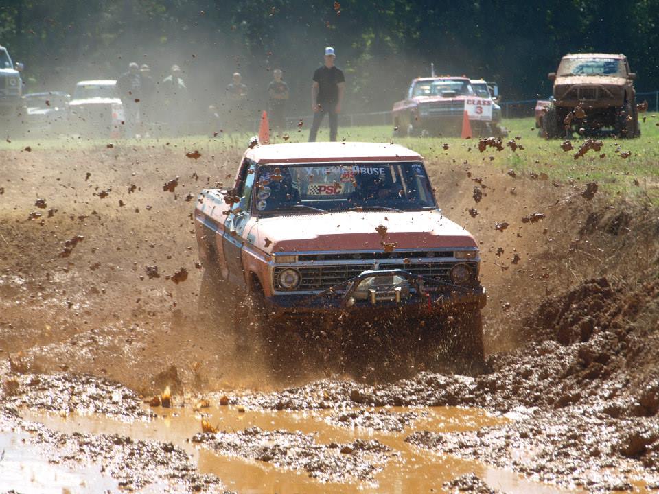 Orange Ford pickup truck splashing through mud pit, people watching in background