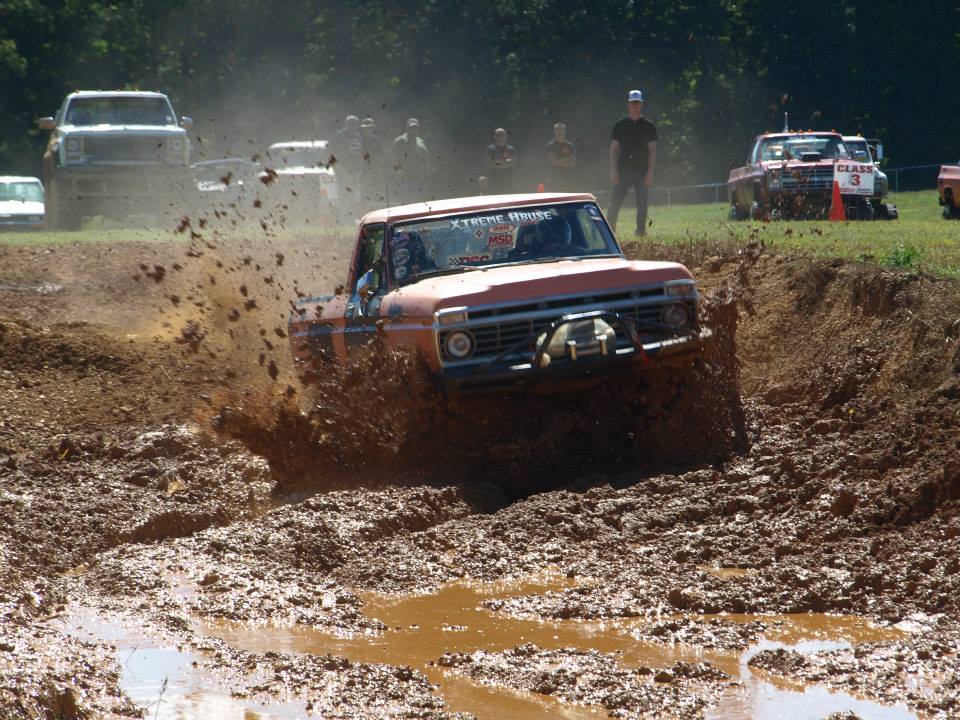 Orange pickup truck with a bull bar driving through a mud pit. Other trucks and people are visible in the background