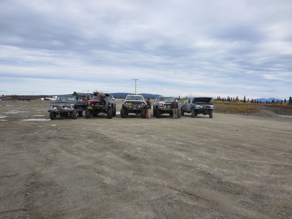 Five modified trucks and off-road vehicles parked in a dirt lot under a cloudy sky