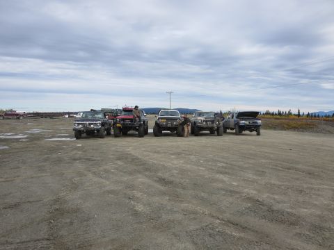 1990 Ford F-250 - A group of modified trucks are gathered in an outdoor lot with a wooded area in the background.