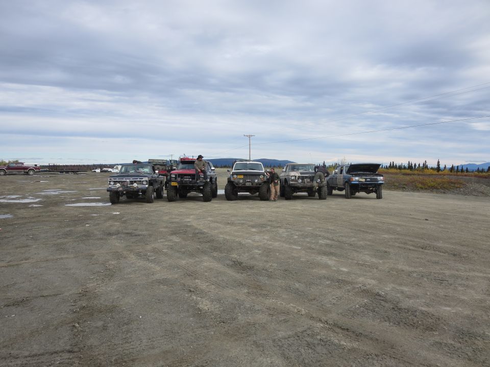 Several lifted trucks parked in a dirt lot under a cloudy sky