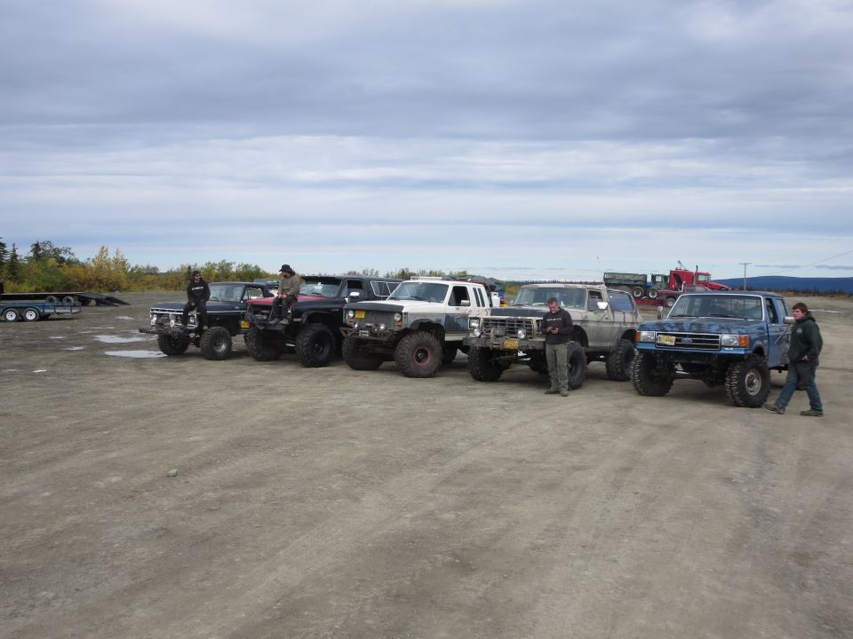 Several lifted pickup trucks parked in a dirt lot with people standing around them