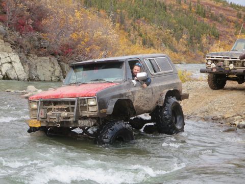 1990 Ford F-250 - A lifted SUV navigates a rocky riverbed, with a second off-road vehicle visible on the bank.