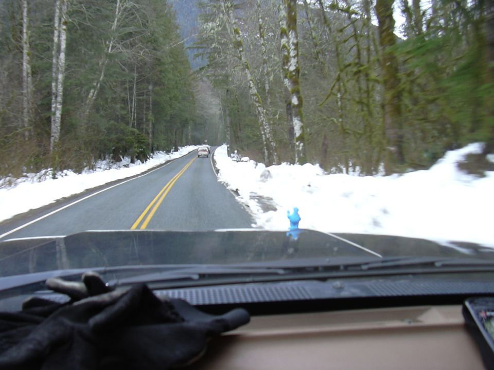 view from inside a vehicle driving on a road with snow on the sides, another car ahead
