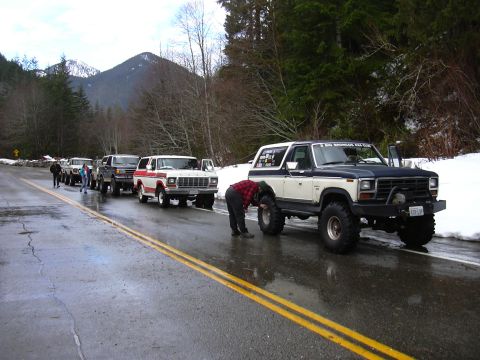 1991 Ford Bronco - A line of Ford Broncos drive on a wet road with snow on the sides.