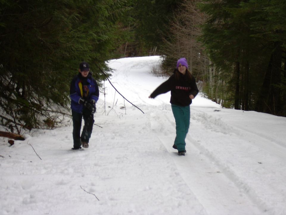 Two people walk on a snow-covered path through a forest. One person wears a blue jacket and a baseball cap, the other wears a dark hoodie and a purple hat