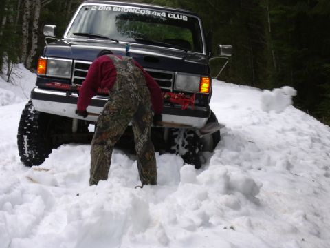 1991 Ford Bronco - A person in camouflage overalls attempts to free a dark blue 1991 Ford Bronco from a snowdrift, with a club sticker visible on the windshie…