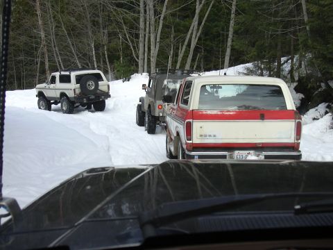 1991 Ford Bronco - A convoy of SUVs, including a red and white Ford Bronco, navigates a snowy road through a forest.