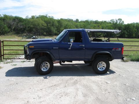 1982 Ford Bronco - This lifted Ford Bronco is parked on a dirt road with a fence and trees in the background.