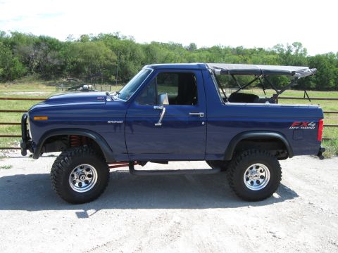 1982 Ford Bronco - A lifted blue Ford Bronco with a removable soft top sits on a gravel surface, with trees and a fence in the background.