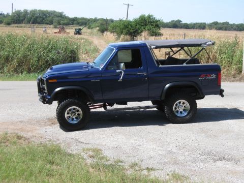 1982 Ford Bronco - A lifted 1982 Ford Bronco with a removable soft top sits parked on the side of a road, with agricultural fields visible behind it.