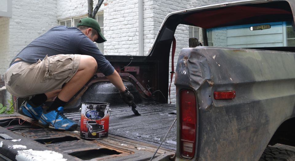 Man crouches in bed of 1979 Ford Bronco, painting the surface with a brush. Paint can visible