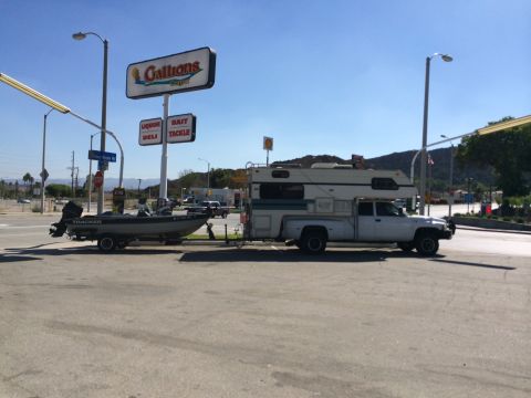 1997 Dodge 3500 - A white pickup truck with a camper shell is parked, towing a boat on a trailer, with a store sign in the background.