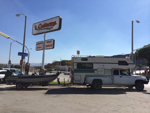 1997 Dodge 3500 - A white truck with a camper and boat trailer is parked near a Gallions store.