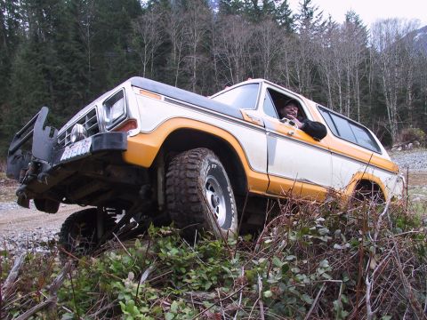 1991 Ford Bronco - A man drives a lifted Ford Bronco through rough terrain, navigating over brush and uneven ground.