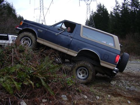 1991 Ford Bronco - A blue and tan 1991 Ford Bronco navigates an off-road trail, ascending a steep, muddy incline.