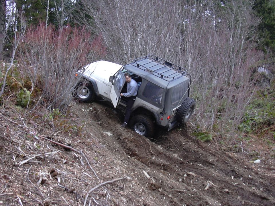 Jeep Wrangler with roof rack stuck on a muddy incline, driver's door open