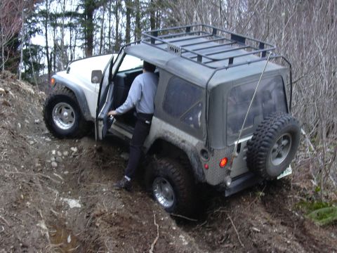 1991 Ford Bronco - A person exits a Jeep Wrangler TJ on a muddy, wooded trail, with the vehicle angled on uneven terrain.