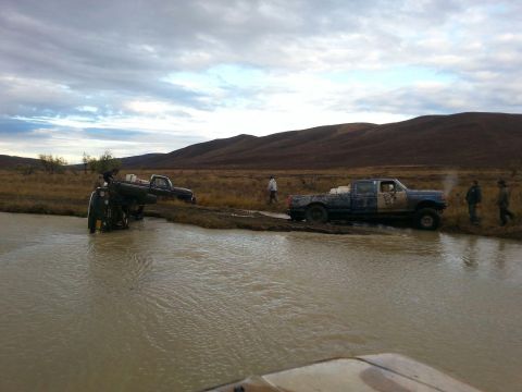 1990 Ford F-250 - Several trucks are stuck in a muddy body of water with hills in the background.