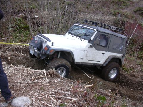 1991 Ford Bronco - A white Jeep Wrangler navigates a challenging off-road terrain, with one wheel stuck in the mud.