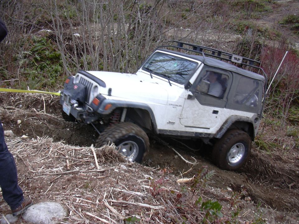 White Jeep Wrangler struggling on muddy incline, front wheel deep in dirt