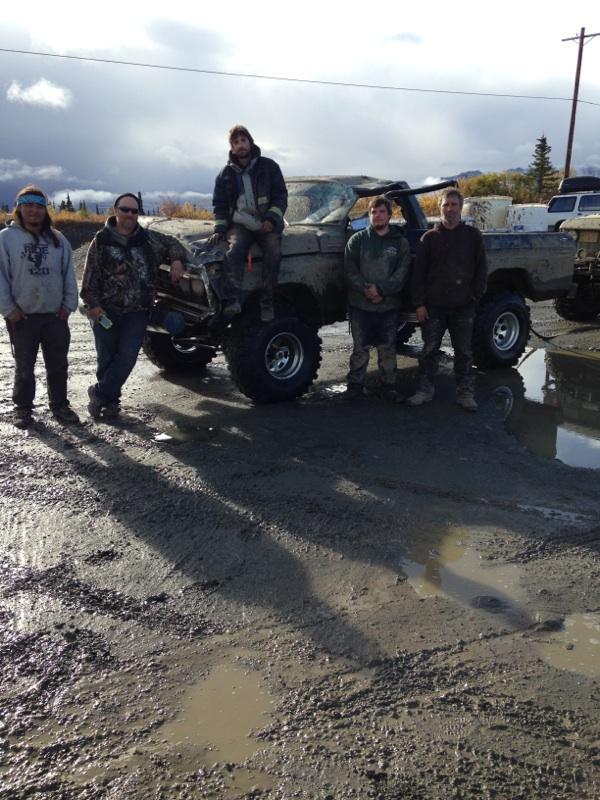 Five men standing around a heavily modified pickup truck in a muddy area. Another vehicle is visible to the right