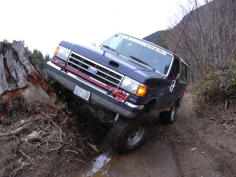 1991 Ford Bronco - A dark blue Ford Bronco navigates a muddy trail, with a large tree stump to its left.