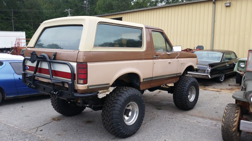 Lifted 1987 Ford Bronco, two-tone brown and tan, with aftermarket rear bumper and spare tire carrier. Blue car and black sedan visible behind