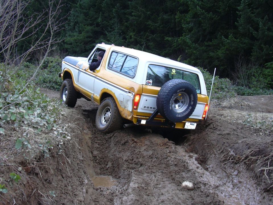 Rear view of a white and yellow 1978 Ford Bronco driving on a muddy off-road trail