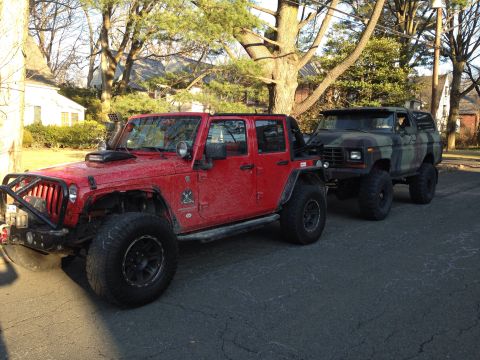 1979 Ford Bronco - A 1979 Ford Bronco in camouflage paint is parked behind a red Jeep Wrangler on a paved road.