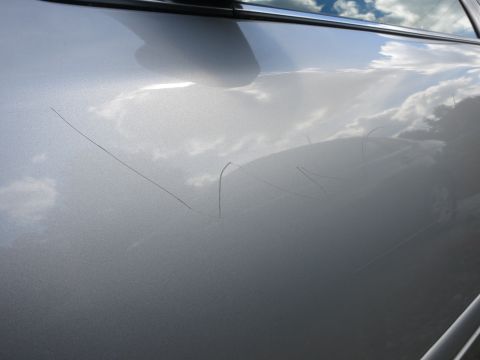 1991 Ford LTD Crown Victoria - The side of a silver car shows multiple long scratches on its paintwork, with reflections of the sky and clouds.