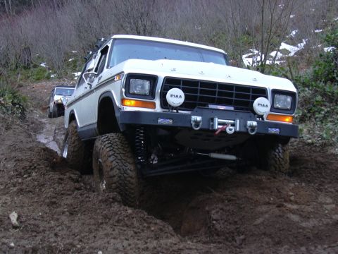 1991 Ford Bronco - A white Ford Bronco navigates a challenging muddy off-road course, with another vehicle following behind.