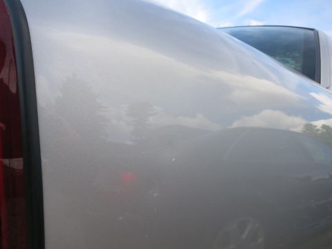 1991 Ford LTD Crown Victoria - Close-up of a silver car's side panel, reflecting the sky and surrounding trees. Another vehicle is partially visible in the background.