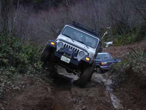 1991 Ford Bronco - Two off-road vehicles navigate a challenging muddy trail with dense vegetation on the sides.