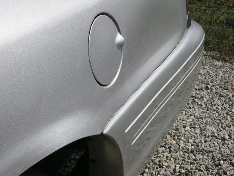 Silver car's rear quarter panel showing the fuel door and a pinstripe trim. The car is parked on a gravel surface