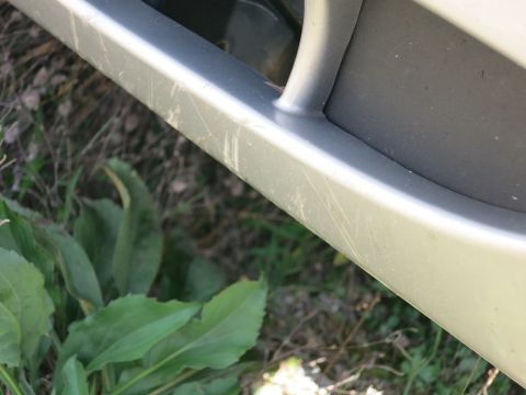 1991 Ford LTD Crown Victoria - A close-up of a scratched silver car bumper, with green foliage visible below.