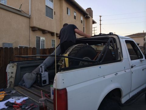 1990 Ford Bronco - A person works on the soft top of a white pickup truck with a spare tire and roll cage visible in the bed.
