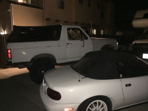 1990 Ford Bronco - A white Ford Bronco with a black soft top is parked next to a white convertible.