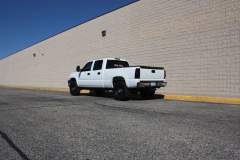 2006 Chevrolet Silverado 2500 - A white pickup truck is parked on an asphalt surface next to a beige brick wall under a clear blue sky.