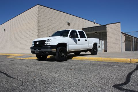 2006 Chevrolet Silverado 2500 - A white 2006 Chevrolet Silverado 2500 pickup truck is parked on asphalt in front of a building with a yellow curb.