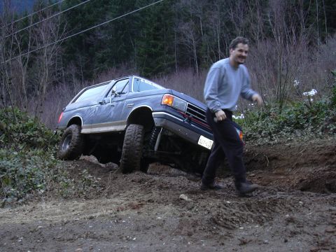 1991 Ford Bronco - A man walks beside a lifted Ford Bronco as it navigates a steep, muddy incline.